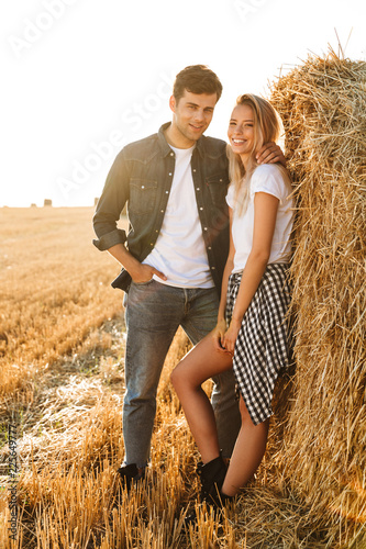 Full length photo of beautiful couple man and woman walking on field after harvesting, and standing near big haystack