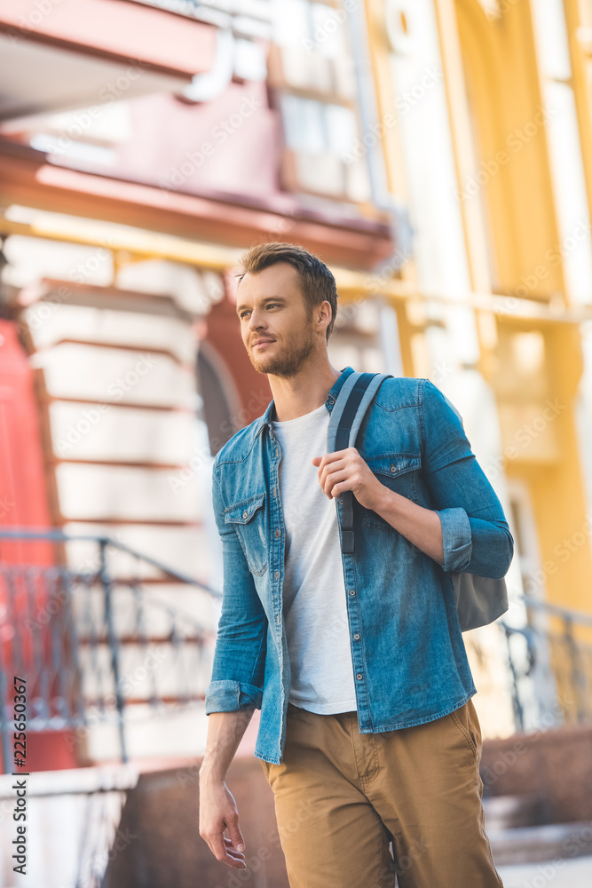 Fototapeta premium attractive young tourist with backpack walking by street and looking away