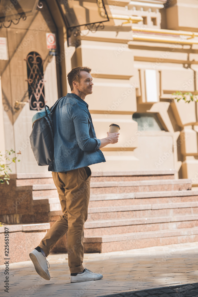 casual young man with paper cup of coffee and backpack walking by street and looking away