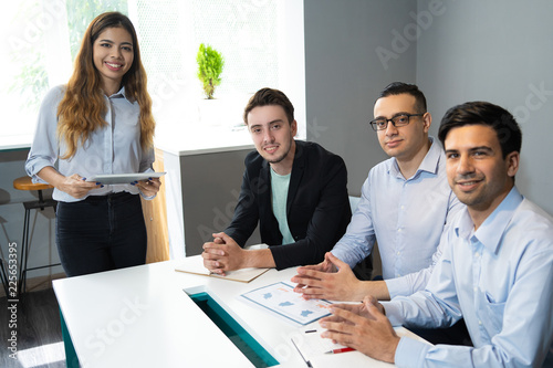 Portrait of successful young business team with female leader. Businessmen sitting at table and standing woman looking at camera and smiling in office. Woman leader concept