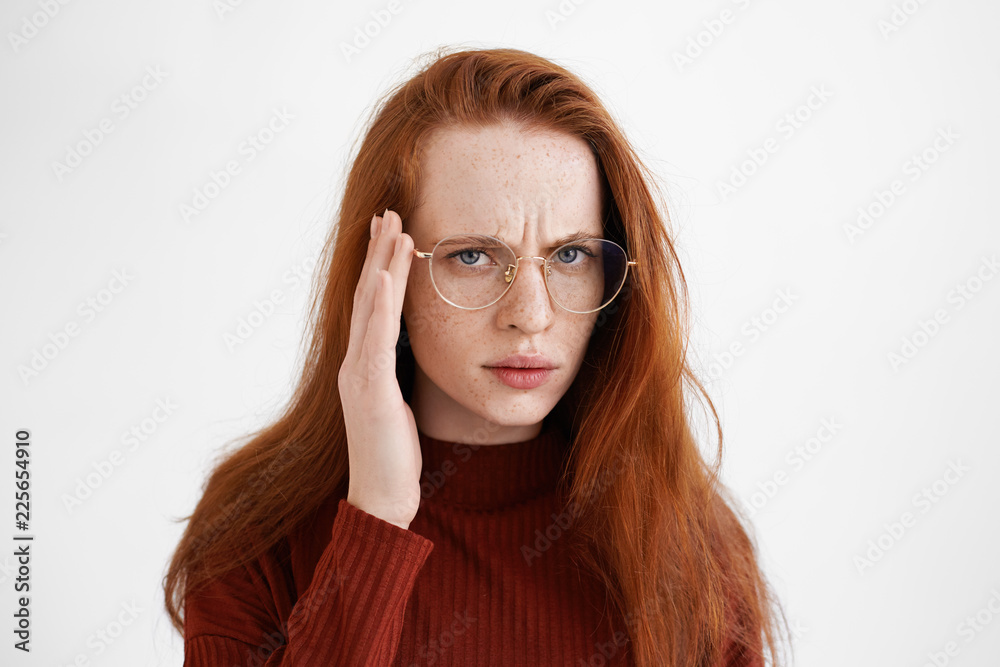 Isolated sudio shot of displeased young Caucasian woman with ginger hair touching rim of her eyeglasses, frowning eyebrows and staring at camera suspiciously, having distustful facial expression Isolated sudio shot of displeased young Caucasian woman with ginger hair touching rim of her eyeglasses, frowning eyebrows and staring at camera suspiciously, having distustful facial expression
