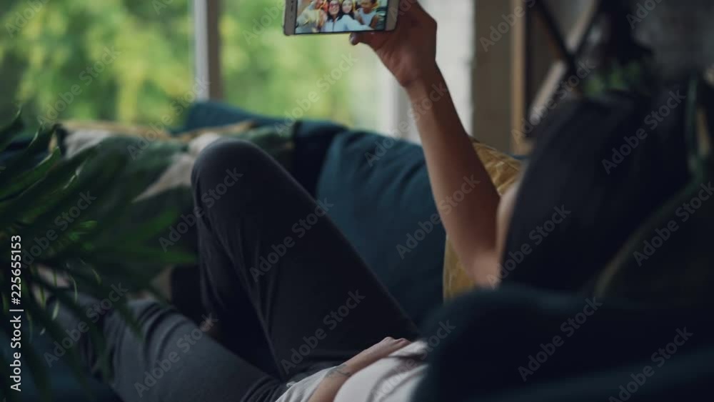 Rear view of brunette making online video call holding smartphone looking at screen talking and gesturing indoors at home on comfy sofa. Friendship and technology concept.