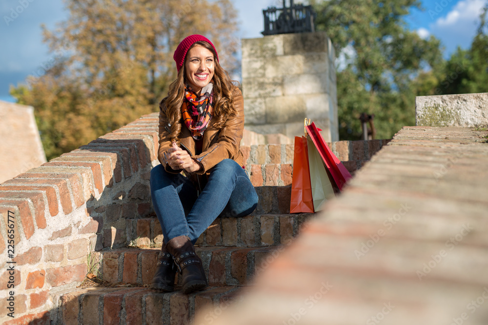 Fototapeta premium Beautiful young woman sitting on stairs outside while drinking a coffee after shopping