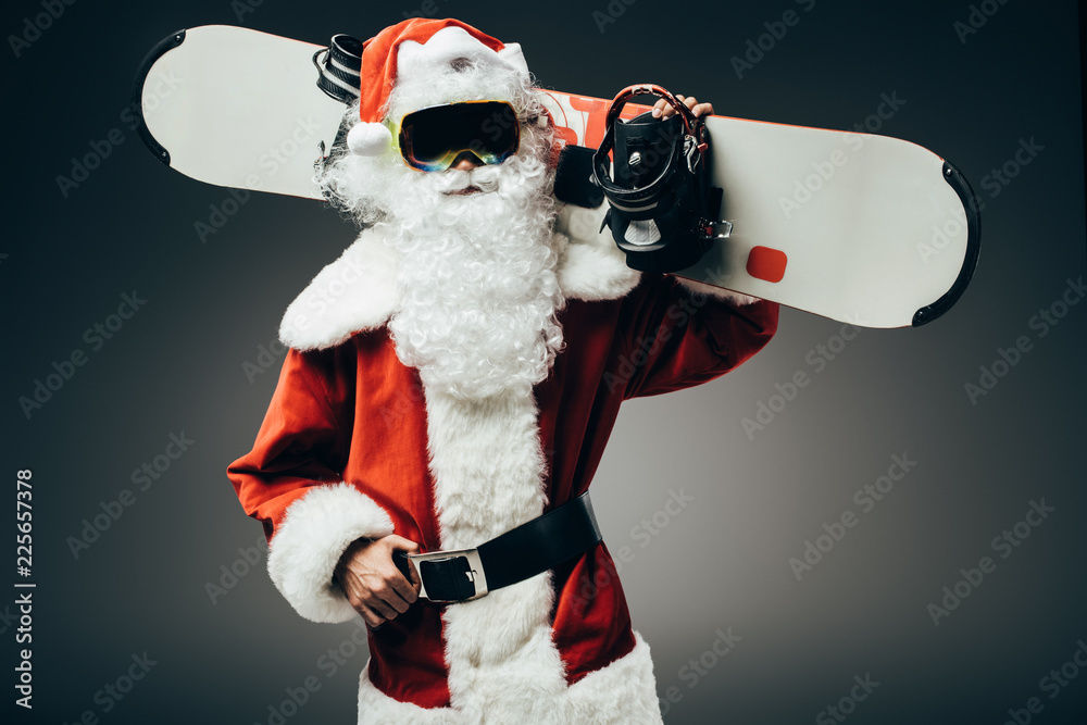 Fototapeta poważny Święty Mikołaj w masce narciarskiej stojący z snowboard na ramieniu na białym tle na szarym tle