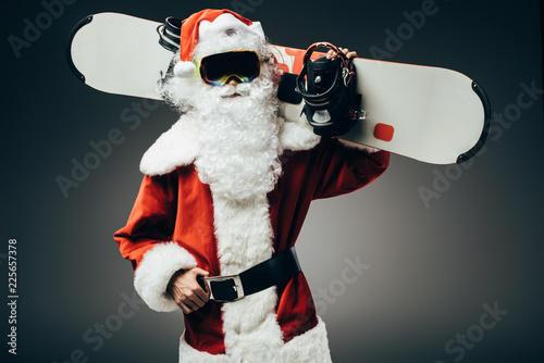 serious santa claus in ski mask standing with snowboard over shoulder isolated on grey background
