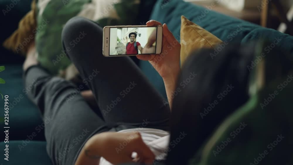 Young woman with long dark hair is making online video call holding smartphone and gesturing talking to male friend lying on sofa in house. Youth and communication concept. Young woman with long dark hair is making online video call holding smartphone and gesturing talking to male friend lying on sofa in house. Youth and communication concept.