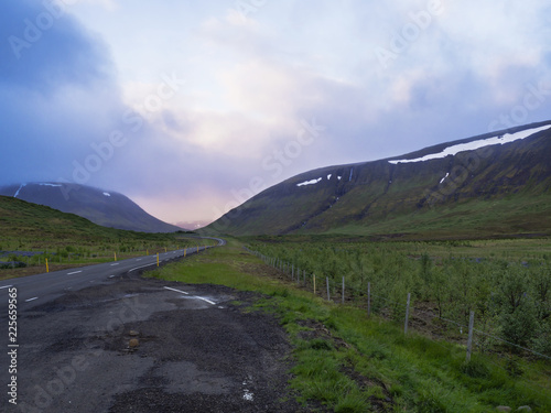Asphalt road curve through empty northern landscape with green grass colorful hills and sunset dramatic sky, way to the mountains in Iceland western highlands