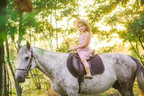 Little cute girl with light curly hair in a straw hat riding a horse at sunset on a sunny warm autumn day 