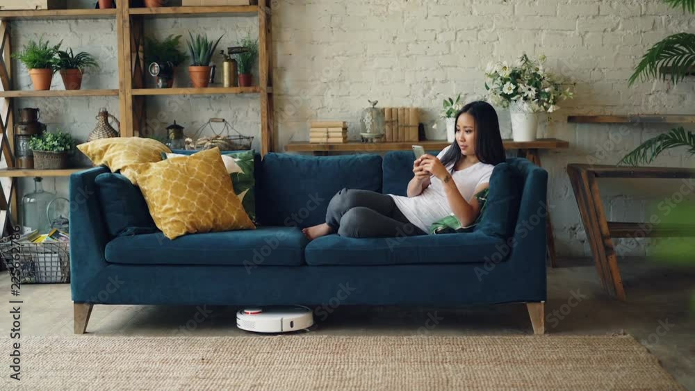 Attractive Asian girl is enjoying modern technology using smartphone and robotic vacuum cleaner sitting on sofa at home. Innovations and interiors concept. Attractive Asian girl is enjoying modern technology using smartphone and robotic vacuum cleaner sitting on sofa at home. Innovations and interiors concept.