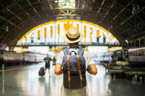 Young man traveler with backpack waiting for train, Asian backpacker with hat standing on railway platform at Bangkok train station. Holiday, journey, trip and summer Travel concept
