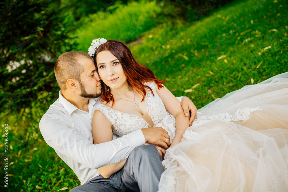 Naklejka premium wedding, beautiful young bride lying together with groom in love on green grass kissing, park summer outdoor. Newlyweds wedding photoshoot.