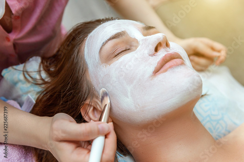 Close-up of young beauty brunette woman getting face treatment with white nourishing creme in spa salon. Face massage. Spa skin and body care. Skincare cleansing cosmetic spa relax concept