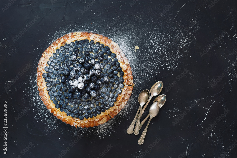 Delicious homemade blueberry almond tart with fresh blueberries and bunch of teaspoons beside. Top  view, blank space, dark toned image - obrazy, fototapety, plakaty 