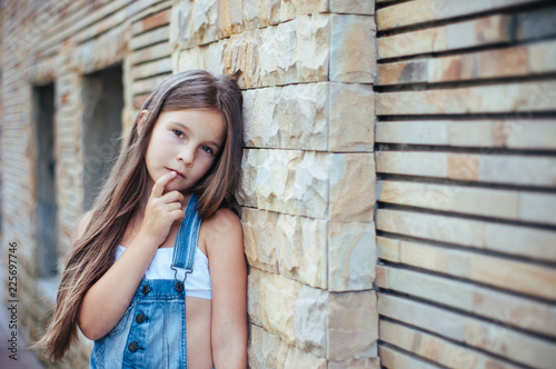Model little beautiful girl with long hair, street fashion near the wall on a summer
