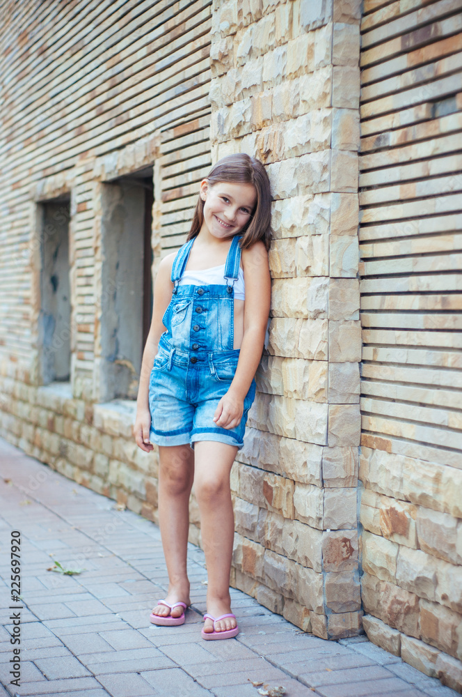 Model little beautiful girl with long hair, street fashion near the wall on a summer