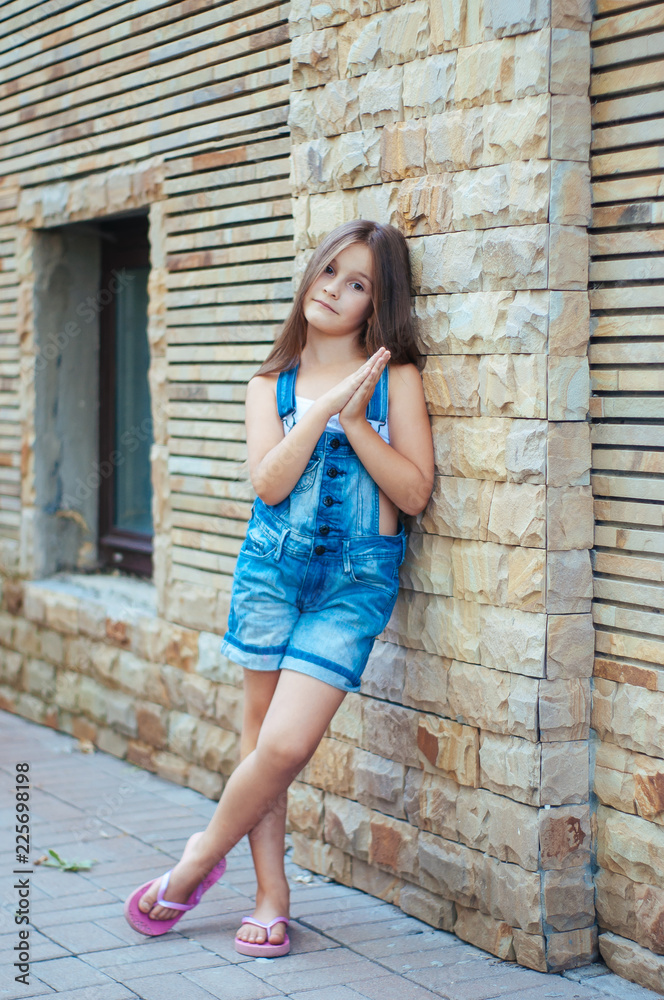 Model little beautiful girl with long hair, street fashion near the wall on a summer