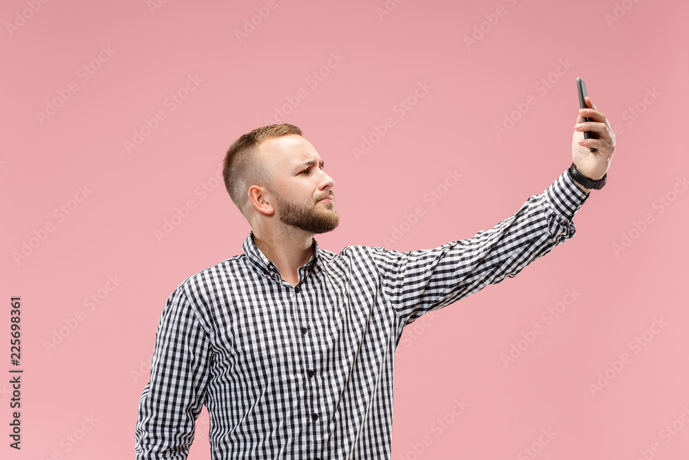 Portrait of attractive serious young man taking a selfie with his smartphone. Isolated on pink studio background. Human emotions, facial expression concept. Trendy colors Portrait of attractive serious young man taking a selfie with his smartphone. Isolated on pink studio background. Human emotions, facial expression concept. Trendy colors