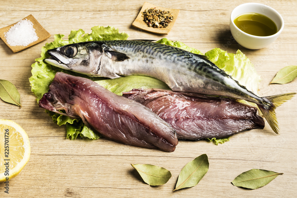 Mackerel fresh fish on lettuce leaves next to bay leaves, a few pieces of lemon, a bowl of oil and some spices on a wooden table Mackerel fresh fish on lettuce leaves next to bay leaves, a few pieces of lemon, a bowl of oil and some spices on a wooden table