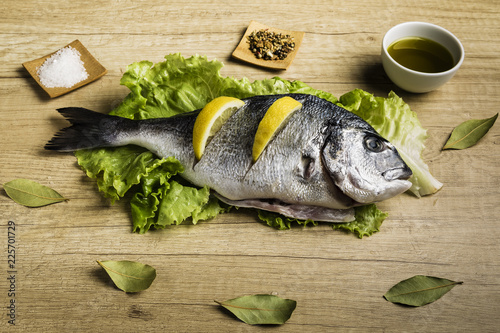 Dorada fresh fish on lettuce leaves next to some bay leaves, some pieces of lemon, a bowl of oil and some spices on a wooden table