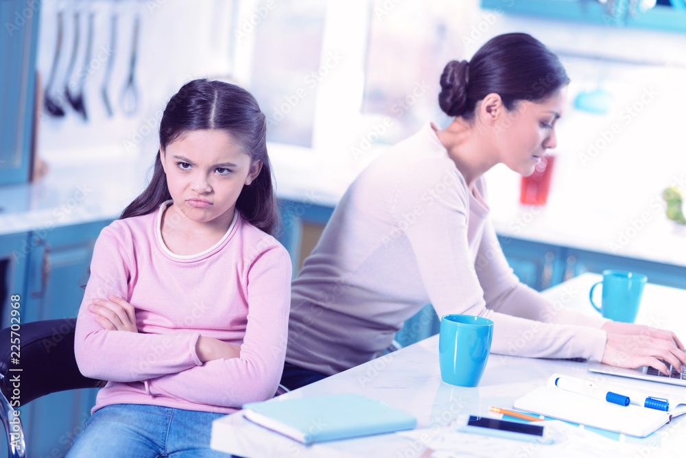 Feeling offended. Sad little girl sitting next to her busy mother with her arms crossed and feeling offended Feeling offended. Sad little girl sitting next to her busy mother with her arms crossed and feeling offended
