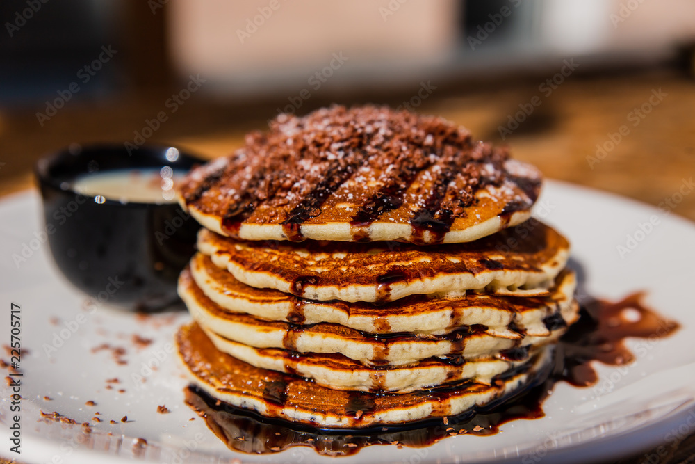A horizontal photo of pancakes topped with chocolate sauce, powdered sugar and chopped chocolate, served on a white plate with sweetened condensed milk A horizontal photo of pancakes topped with chocolate sauce, powdered sugar and chopped chocolate, served on a white plate with sweetened condensed milk