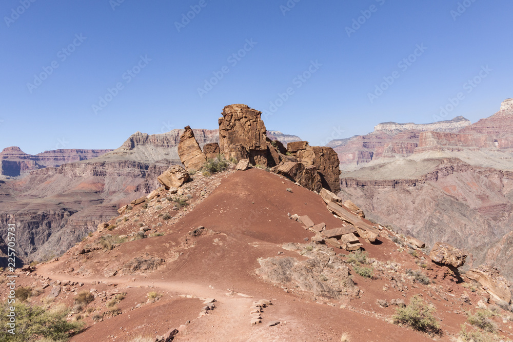 Naklejka premium Beautiful rock boulders on a hill of red sand on a hiking trail down the Grand Canyon with a view of the famous rock formations on the background.