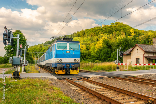 The blue electric train arriving at the crossing with the barriers. A train running through the green valley. Rail transport in the Czech Republic. A sunny day on the railroad