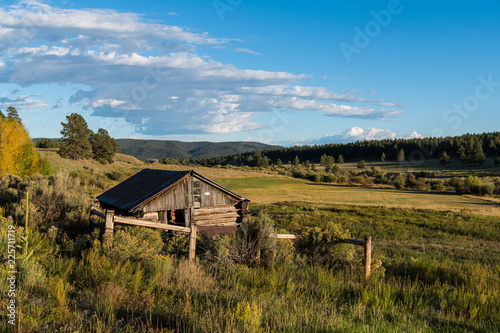 Old rustic log cabin overlooking a landscape of ranchland, fields, forest, and hills in northern New Mexico in late afternoon sunlight