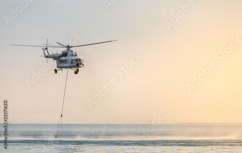 A helicopter with a red basket descends over the sea to scoop water against the background of the dawn orange sky and the silhouette of the city in the distance