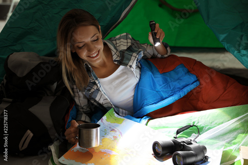 Young woman in sleeping bag with map and flashlight inside of tent
