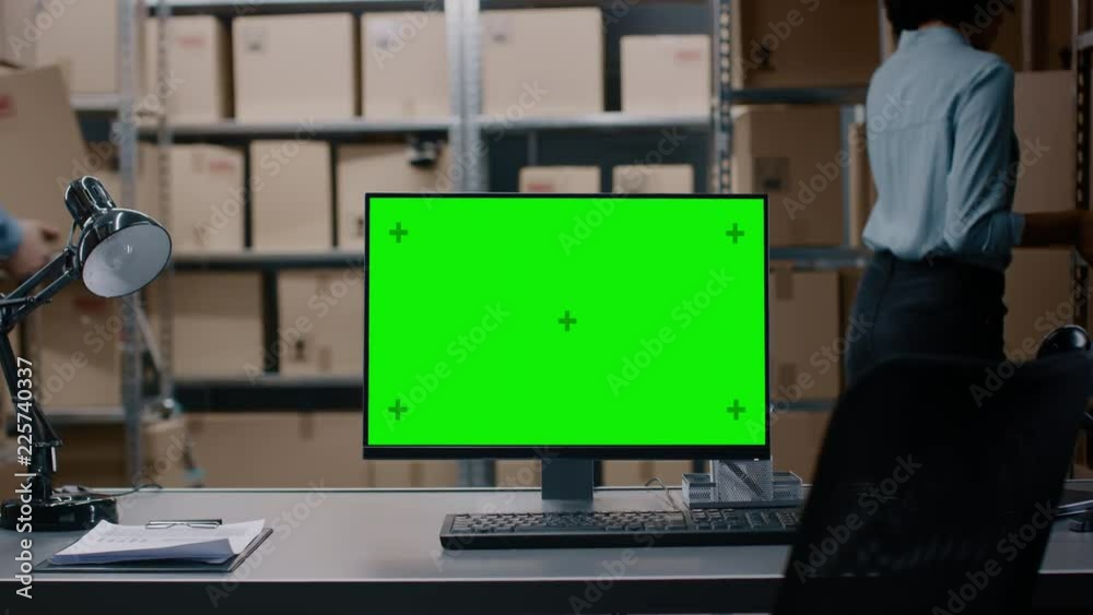 In the Warehouse Computer with Green Mock-up Screen Display Standing on Desktop. In the Background Rows of Shelves Filled With Cardboard Boxes and Parcels with Products Ready to be Shipped.