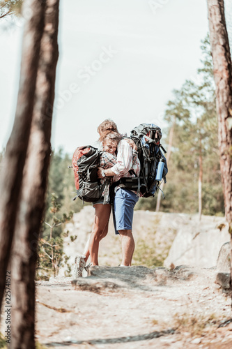 Hugs and hiking. Tender curly dark-haired woman with heavy backpack hugging her strong bearded man while hiking