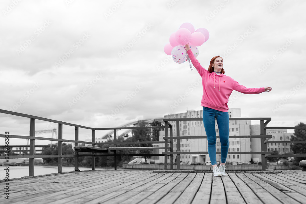 Little party. Happy emotional young girl smiling while standing on the pier and holding a bunch of pink balloons for the party