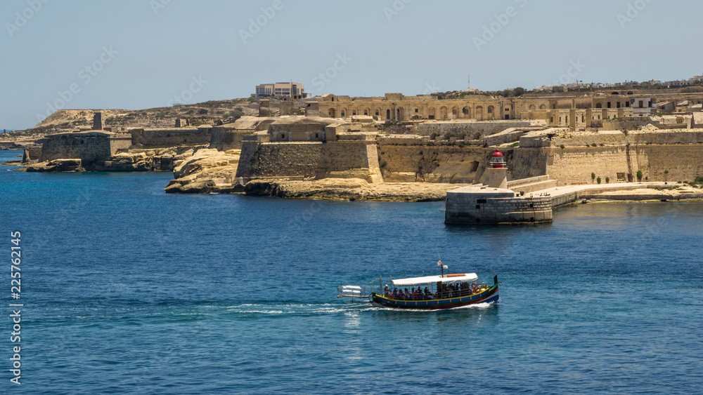 A traditional Maltese boat called a Luzzu passes the Fort Ricasoli and the Ricasoli Breakwater lighthouse at the entrance to the Grand Harbour, Malta.