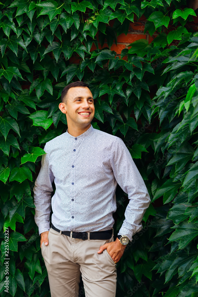 sincere smile of a guy who holds his hands in his pockets against the background of a building on which the facade of a plant with green leaves sincere smile of a guy who holds his hands in his pockets against the background of a building on which the facade of a plant with green leaves