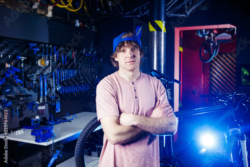 Small business and bicycle transport service. Portrait of a young man in a cap posing against the backdrop of a bicycle workshop and a tool for setting up and repairing © Elizaveta