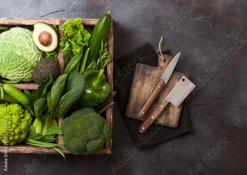 Assorted green toned raw organic vegetables in wooden box on dark background. Avocado, cabbage, cauliflower and cucumber with trimmed and mung beans and chopping board with knife. photo