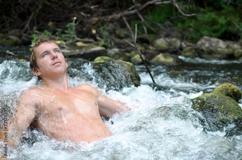 young handsome man lies in a waterfall formed by a rapids river and the water flows around his athletic body