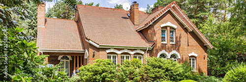 Canvas Print A red brick English style classic house with a steep roof and large windows surrounded by trees and green plants