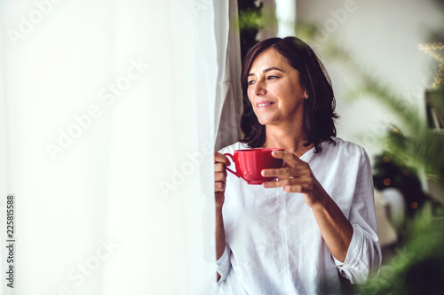 Tableau sur toile A senior woman with a cup standing by the window at home at Christmas time
