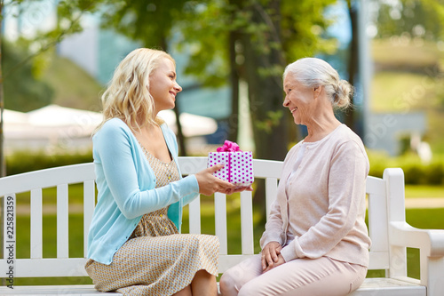 family, holidays and people concept - happy smiling young daughter giving present to her senior mother sitting on park bench