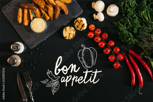 top view of tasty baked potatoes with sauce, spices and vegetables on black with "bon appetit" lettering