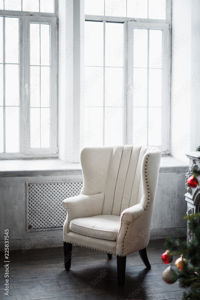 White large cozy chair in a room with gray walls and a Christmas tree in the foreground blurred. Selective focus. White large cozy chair in a room with gray walls and a Christmas tree in the foreground blurred. Selective focus.