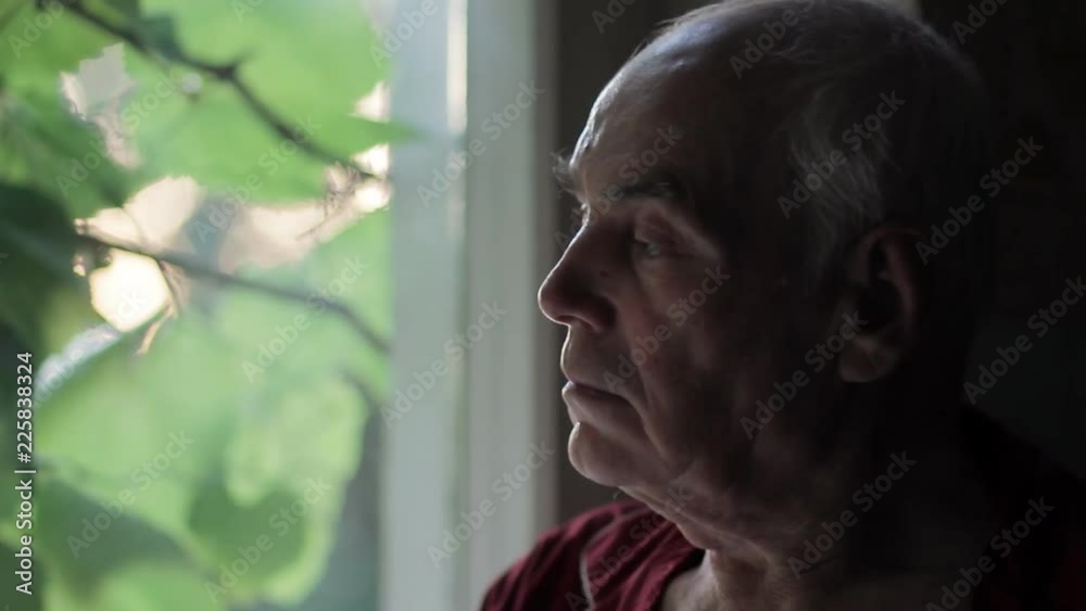 Portrait of sad lonely elderly man looking out of the window at sunset. Loneliness and depression in old age.
