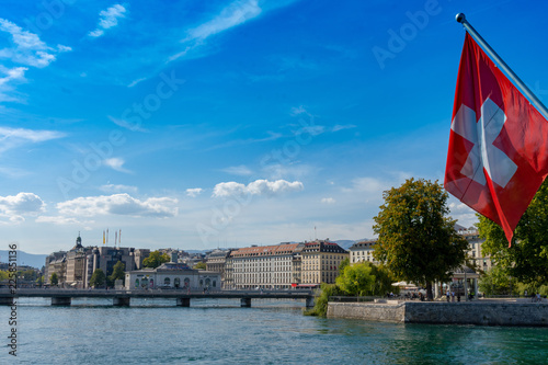Swiss flag in front of the geneve skyline, cathedrale saint-pierre geneve, yellow boat and a wonderful blue sky as seen from the pont du mont blanc bridge of Geneva, Switzerland photo