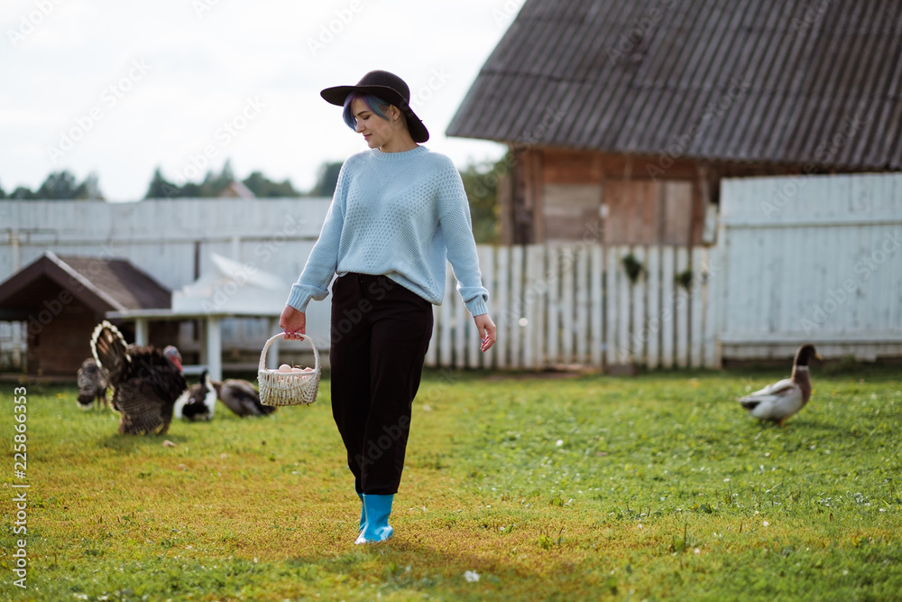 Fototapeta premium Young beautiful girl in a village holding a basket with chicken eggs. Autumn day, green grass, on the background of a farm. A woman is smiling in a sweater and a black hat.