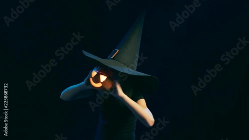 Happy playful child girl wearing black witch hat holding jack-o'-lantern in front of face dancing then lowering pumking smiling at camera, over black background photo