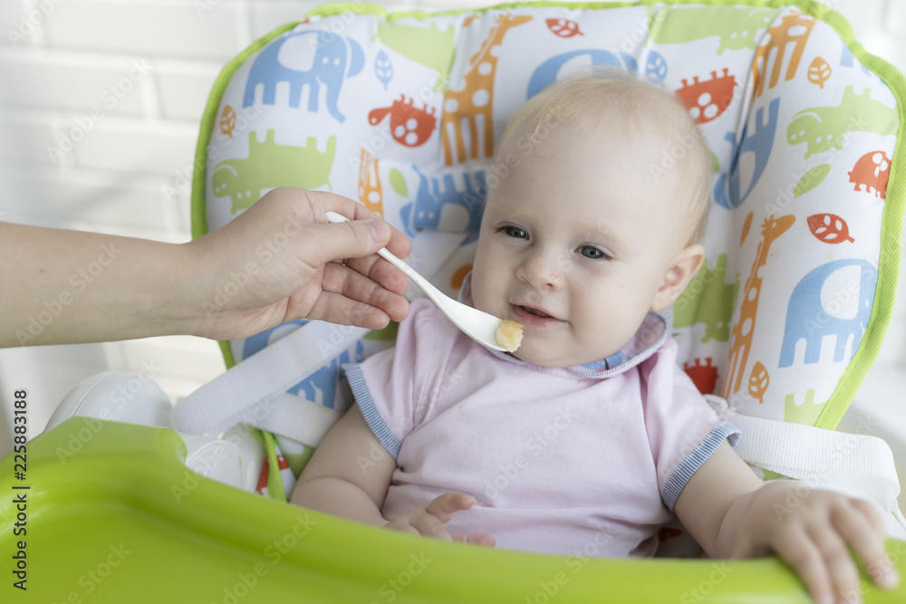 mother feeds her little daughter sitting in a baby chair. The mother feeds a small baby delicious food with a spoon. The baby enjoys while eating a meal. mother feeds her little daughter sitting in a baby chair. The mother feeds a small baby delicious food with a spoon. The baby enjoys while eating a meal.