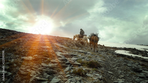 A rider with two horses walks along the path to the mountains. Bags with provisions on horses. The rays of the sun look out from behind the top. Soft blur effect. photo