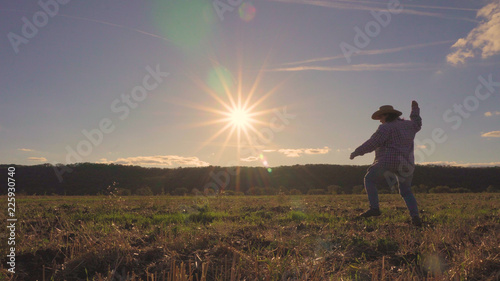 A woman walks across the field and leans to the ground. On the background of the sunset. Warm weather. Straw hat on his head. Shirt's on. Cleaned field. A rustic appearance. Blue sky with clouds. © Evgeniy Trofimenko